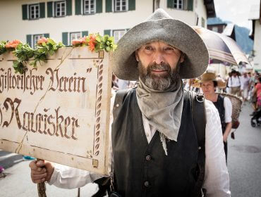 Ein Mann in traditioneller Kleidung hält ein geschmücktes Schild bei einem Freiluftfest oder einer Parade in einem Dorf.