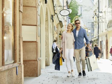 Ein lächelndes Paar geht mit mehreren Einkaufstaschen durch eine sonnige Einkaufsstraße.