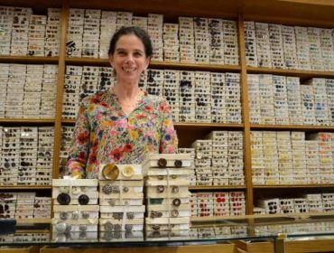 Eine Frau steht hinter einem Tresen, vor sich Kisten mit Knöpfen, hinter sich Regale mit Knöpfen.