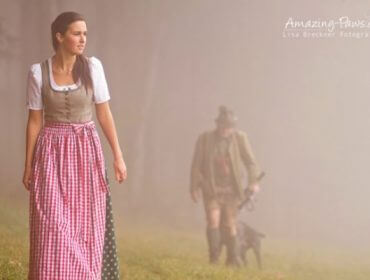 Frau in traditioneller österreichischer Tracht geht in einem nebligen Feld spazieren; Mann und Hund folgen im Hintergrund.