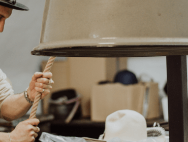 Eine Person, die einen Hut trägt, arbeitet mit einem Seil in einer Werkstatt, umgeben von Hüten und Werkzeugen.