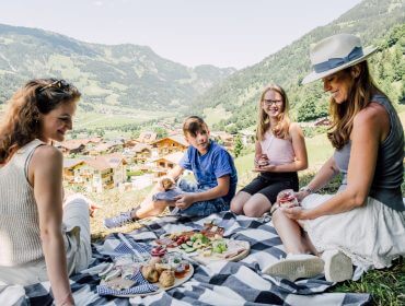 Genussreise Gastein Vier Personen machen ein Picknick auf einer Decke mit Blick auf ein österreichisches Dorf und die Berge an einem sonnigen Tag.