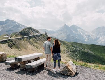Genussreise Nationalpark Hohe Tauern & Großglockner Hochalpenstraße Ein Paar steht auf einer Bank an einem Aussichtspunkt in den Bergen und blickt auf schneebedeckte Gipfel und grüne Hügel.