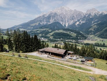 Eine von Bäumen umgebene Berghütte, mit entfernten Dörfern und großen Bergen im Hintergrund.