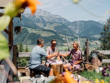 Sinnlehenalm Drei Personen sitzen draußen an einem Tisch und stoßen mit Getränken an, während im Hintergrund Berge und Grünflächen zu sehen sind.