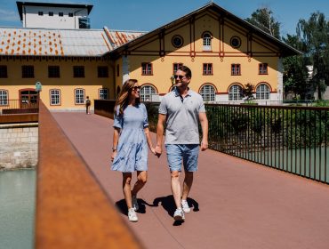 Ein Paar, das sich an den Händen hält und an einem sonnigen Tag auf einer Brücke neben gelben Gebäuden spazieren geht.