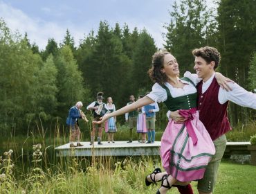 Ein lächelndes Paar in traditioneller Kleidung tanzt im Freien, während hinter ihnen Menschen in ähnlicher Kleidung auf einem Holzsteg stehen.