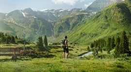 Ein Wanderer mit einem Rucksack steht in einem grünen Tal und blickt auf hohe Berge unter einem klaren Himmel.