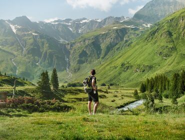 Ein Wanderer mit einem Rucksack steht in einem grünen Tal und blickt auf hohe Berge unter einem klaren Himmel.