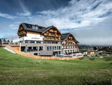 Ein modernes Alpenhotel mit Holzbalkonen liegt an einem grünen Hügel unter einem teilweise bewölkten Himmel.