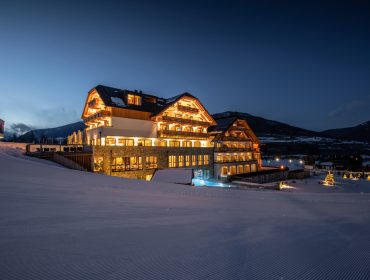Ein warm beleuchtetes Alpenhotel an einem verschneiten Berghang in der Abenddämmerung, mit Bergen und einem kleinen Dorf im Hintergrund.