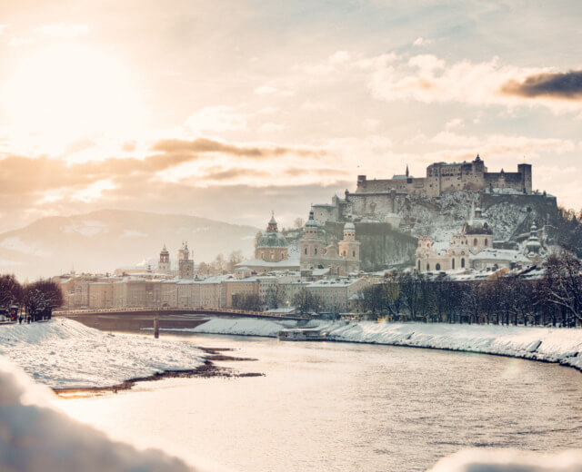 Verschneite Stadt Salzburg Verschneite Stadtlandschaft von Salzburg, Österreich, mit einem Fluss, historischen Gebäuden und einer Festung auf einem Hügel bei Sonnenuntergang.