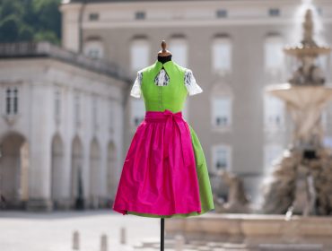 Grünes und rosafarbenes traditionelles Dirndlkleid an einer Schaufensterpuppe vor einem verschwommenen europäischen Brunnen und Gebäude.