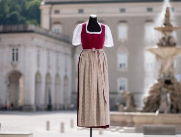 Eine Schaufensterpuppe zeigt ein traditionelles österreichisches Dirndlkleid auf einem europäischen Stadtplatz in der Nähe eines Steinbrunnens.