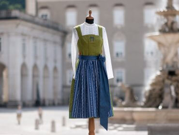 Ein traditionelles österreichisches Dirndlkleid an einer Schaufensterpuppe steht im Freien vor einem verschwommenen historischen Gebäude und einem Brunnen.