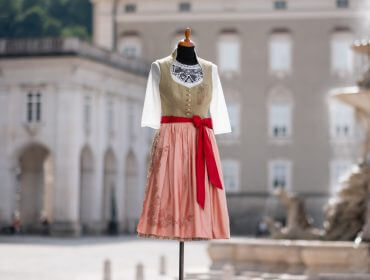 Schaufensterpuppe, die ein traditionelles Dirndlkleid mit roter Schärpe auf einem sonnenbeschienenen europäischen Platz trägt.