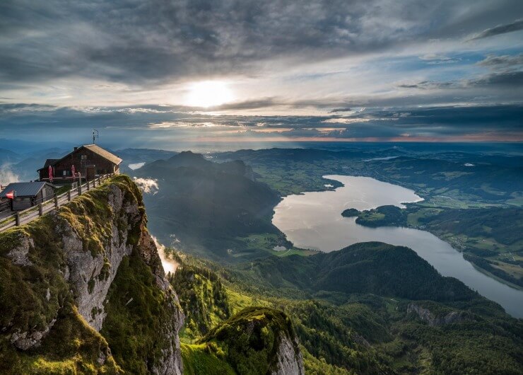 Eine Berghütte auf einer Klippe überblickt einen See und grüne Täler bei Sonnenaufgang unter einem teilweise bewölkten Himmel.