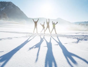 Foto Salzkammergut Tourismus/Karl Steinegger: Winter Fun im Salzkammergut Vier Menschen springen auf einem verschneiten Feld mit langen Schatten, Bergen und Sonne im Hintergrund.