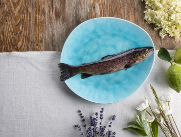 Ein ganzer Fisch auf einem blauen Teller, umgeben von Zitronen, Blumen und Kräutern auf einem rustikalen Tisch.