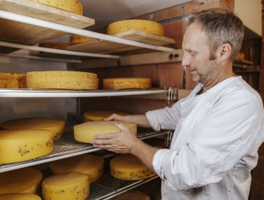 kaese Ein Mann in einem weißen Kittel legt einen runden Laib Käse auf ein Metallgestell in einem Käselagerraum.