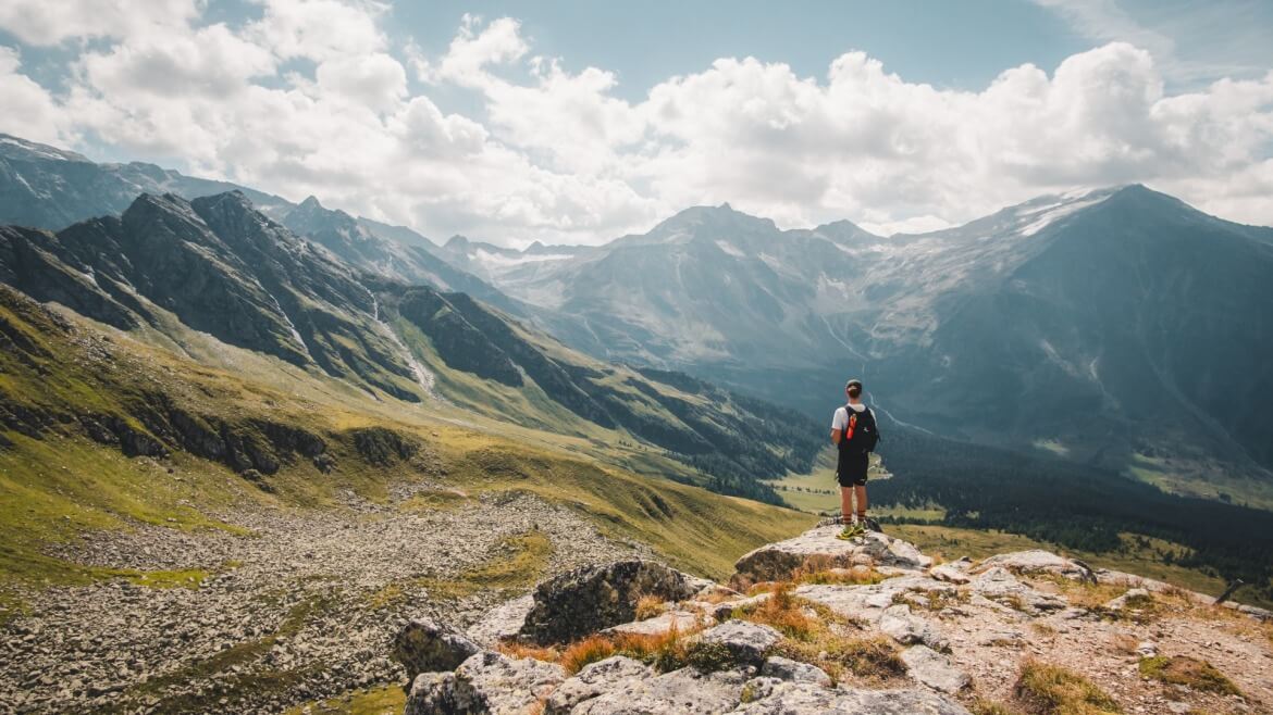 Ein Wanderer steht auf einem Felsvorsprung und überblickt eine weite Berglandschaft bei teilweise bewölktem Himmel.