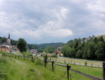 Ein ländliches Dorf mit einer Kirche, Häusern, grünen Hügeln, Bäumen und einem bewölkten Himmel. Ein Holzzaun führt entlang eines Weges.