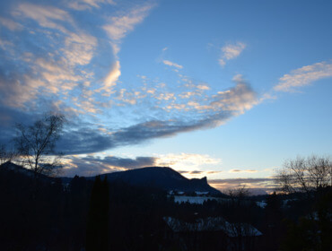 Eine Berglandschaft bei Sonnenuntergang mit dramatischen Wolken und Silhouetten von Bäumen im Vordergrund.