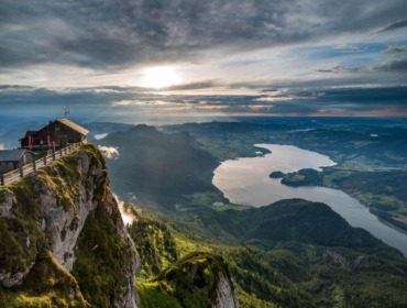 schafberg-himmeslpforte-mondsee-c-wtg Eine Berghütte im Salzkammergut blickt bei Sonnenaufgang unter einem dramatischen Himmel auf einen See und ein üppig grünes Tal.
