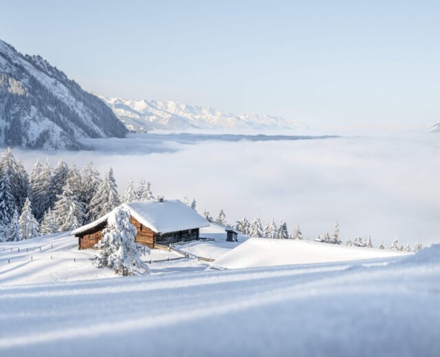 Winterlandschaft © St. Johann in Salzburg Holzhütte in einer verschneiten Berglandschaft über einem Meer aus Wolken.