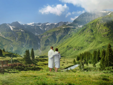 Zwei Personen in weißen Gewändern stehen im Gras und blicken auf grüne Berge und einen kleinen Bach unter blauem Himmel.