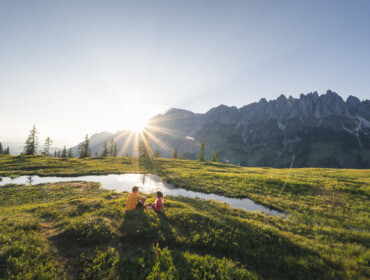 Ein Erwachsener und ein Kind sitzen an einem Teich auf einem grasbewachsenen Hügel und genießen das Wandern von Hütte zu Hütte mit den Bergen im Hintergrund.