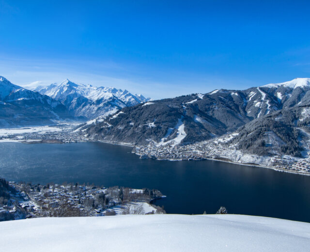 Hauptmotiv Mitterbergblick © Zell am See-Kaprun Tourismus.d237f04a_original Ein schneebedecktes Dorf und ein See, umgeben von Bergen unter einem strahlend blauen Himmel.