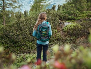 Panorama am Zirbenweg Eine Frau mit Rucksack steht während der Wanderwoche in einer üppigen Berglandschaft und blickt auf ferne Gipfel.