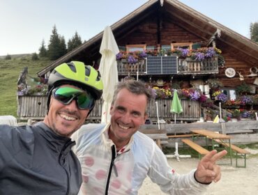 Zwei Männer lächeln vor einer blumengeschmückten Almsommer-Hütte in den Bergen, dazu Felix Gottwald.