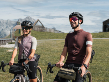 Zwei Radfahrer mit Die-Harte-Helmen lächeln im Freien, Berge und Wiesen im Hintergrund.