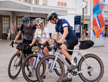 Graveltour SalzburgerLand Drei Radfahrer mit Helmen und Ausrüstung überprüfen ihre Telefone neben ihrem Gravel-Bike an einem modernen Gebäude.