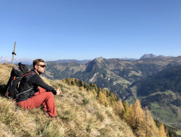 8a923270-fd7e-46d4-bd94-5052f9325c35 Andi Prommegger in roter Hose und Rucksack sitzt auf einem grasbewachsenen Hügel und blickt auf eine malerische Berglandschaft.