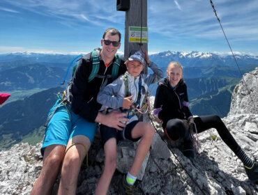 Drei Personen sitzen lächelnd bei einer Gipfelmarkierung auf einem felsigen Berggipfel mit schneebedeckten Gipfeln im Hintergrund.