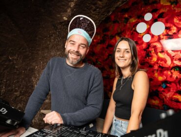 Zwei lächelnde DJs stehen an einem Mischpult in einem farbenfrohen, modernen Raum mit einer leuchtend roten Wandgestaltung.