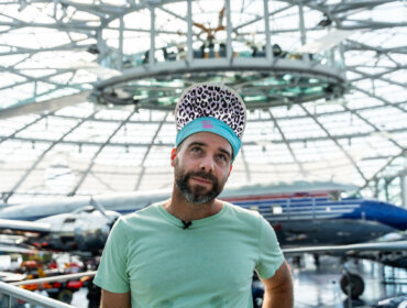 Ein Mann in einem mintgrünen Hemd und einer Mütze mit Leopardenmuster steht in einem Flugzeughangar mit Flugzeugen im Hintergrund.