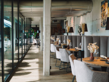 Modernes Restaurantinterieur mit grauen Stühlen, Holztischen und großen Fenstern, die natürliches Licht hereinlassen.