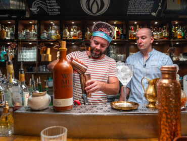 Zwei Barkeeper bereiten hinter einer Bar, umgeben von Flaschen und Gläsern, Getränke zu.