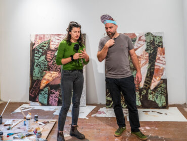 Zwei Künstler stehen vor abstrakten Gemälden und unterhalten sich in einem Atelier, in dem Kunstgegenstände auf dem Boden liegen.