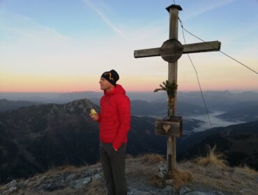 Michael Walchhofer in roter Jacke steht bei Sonnenaufgang an einem Holzkreuz auf einem Berg und hält einen Apfel in der Hand.
