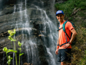 Stefan Kraft steht lächelnd in Wanderkleidung an einem Wasserfall im Grünen, trägt ein orangefarbenes Hemd und eine blaue Mütze.