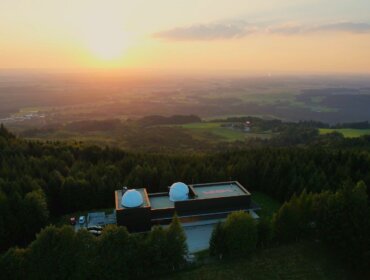 Modernes Observatorium mit zwei Kuppeln, umgeben von Bäumen bei Sonnenuntergang, mit Blick auf eine malerische Landschaft.