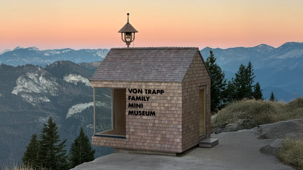 Ein kleines Mini-Museum aus Holz thront bei Sonnenuntergang auf einem Felsvorsprung, im Hintergrund sind ferne Berggipfel zu sehen.