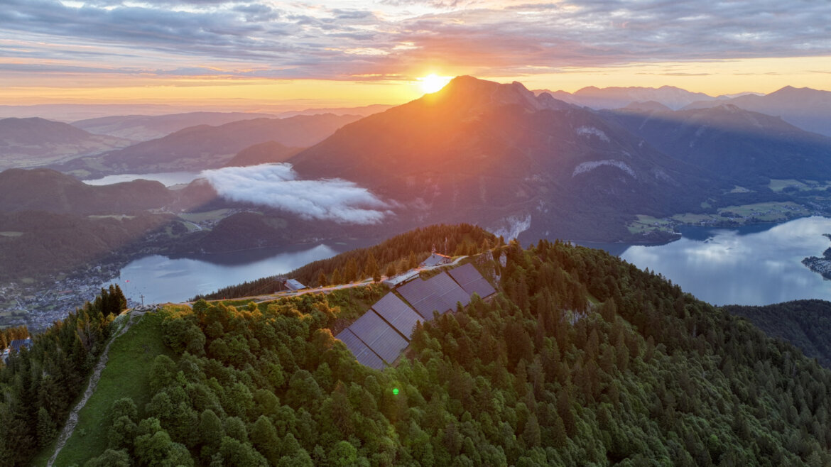 Luftaufnahme einer Berglandschaft bei Sonnenaufgang mit einem See, einem Wald und Sonnenkollektoren im Vordergrund.