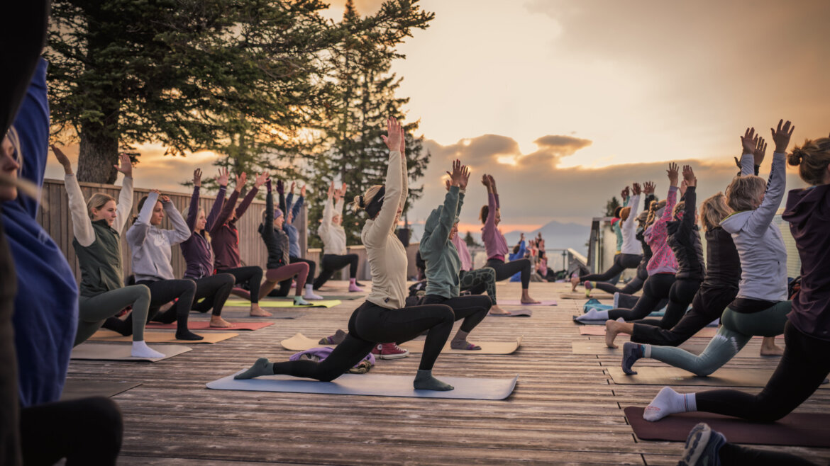 Eine Gruppe von Menschen macht bei Sonnenuntergang auf Matten im Freien Yoga und dehnt sich mit erhobenen Armen.