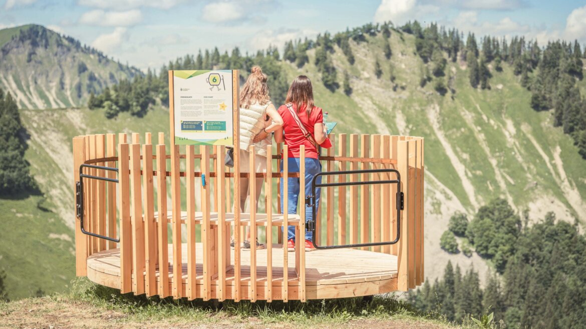 Zwei Frauen stehen an einem sonnigen Tag auf einer hölzernen Aussichtsplattform mit Blick auf eine grüne Berglandschaft. (vergrößerte Ansicht)